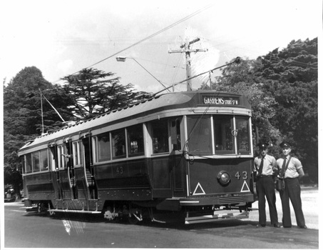 A vintage dark-colored tram numbered 43 with "Gardens Tram" displayed on its front stands at a street, with two uniformed crew members standing beside it.