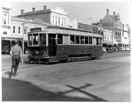 A vintage electric tram with passengers visible through its windows travels down a street lined with multi-story buildings.