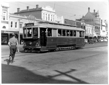 A vintage electric tram with passengers visible through its windows travels down a street lined with multi-story buildings.