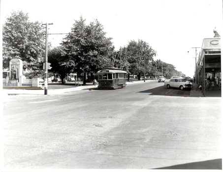 Vintage tram traveling down a tree-lined street in a town with buildings on the right and a monument on the left.
