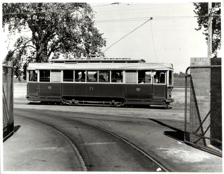 A vintage tramcar labeled "21" travels on a curved track past a chain-link fence under a bright sky.