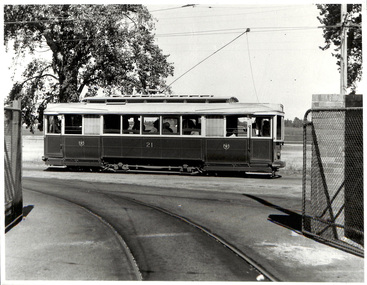 A vintage tramcar labeled "21" travels on a curved track past a chain-link fence under a bright sky.