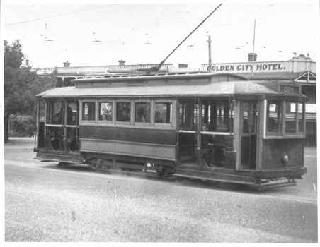 A vintage tram with large windows and overhead electrical wires is positioned on a street in front of a building labeled "GOLDEN CITY HOTEL."