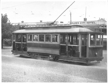 A vintage tram with large windows and overhead electrical wires is positioned on a street in front of a building labeled "GOLDEN CITY HOTEL."