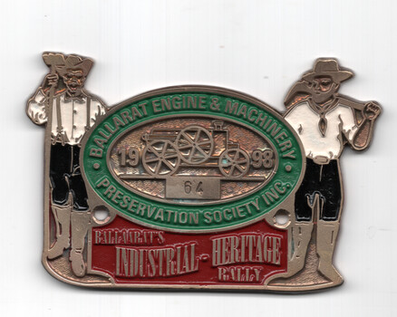 A metal badge for the Ballarat Engine & Machinery Preservation Society Inc. Industrial Heritage Rally, featuring stylized figures holding tools on either side of an oval center with a vintage steam engine illustration and years "1964 - 1998".
