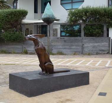 A photograph of a free standing, three dimensional sculpture, cast in bronze of a larger than life dog wearing sunglasses with ears blowing in the wind is sitting under a singular tall tree. Both the dog and tree sit on a large rectangular base. Behind the tree a path and house are visible.