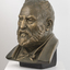 Gilded plaster bust of a balding man with a large round face, creased with age. He wears a trimmed beard, a jacket, a shirt with high neck and bow tie.