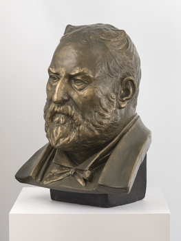 Gilded plaster bust of a balding man with a large round face, creased with age. He wears a trimmed beard, a jacket, a shirt with high neck and bow tie.