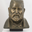 Gilded plaster bust of a balding man with a large round face, creased with age. He wears a trimmed beard, a jacket, a shirt with high neck and bow tie.