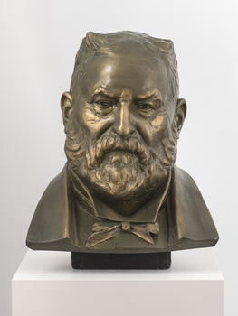 Gilded plaster bust of a balding man with a large round face, creased with age. He wears a trimmed beard, a jacket, a shirt with high neck and bow tie.