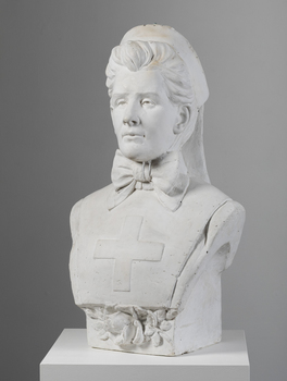 Plaster bust of a woman wearing a stiff, flat garment with medical cross on the chest and a large bow at the neck. Her hair is swept back. She is wearing a nurses cap which has fabric that hangs past her shoulders. A rose is placed under her chest.