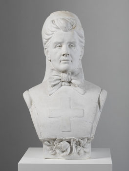Plaster bust of a woman wearing a stiff, flat garment with medical cross on the chest and a large bow at the neck. Her hair is swept back. She is wearing a nurses cap which has fabric that hangs past her shoulders. A rose is placed under her chest.