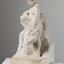 Angled view of an off-white marble statue of a naked woman with a basket of fruit loosely held in her left hand and tipping outwards over her knees. Her right arm leans on a rock. Her feet stand on a rectangular base.