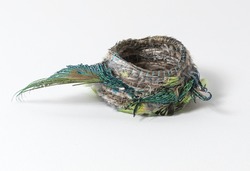 Small woven basket with rows of feathers and green silk thread.  A green peacock feather protrudes from the left side of the basket.