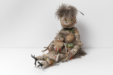 Small woven sculpture of a two figures, a larger female figure and a small child in her lap. Made of woven feathers, cloth, fibre, fur, wood, crow's feet.