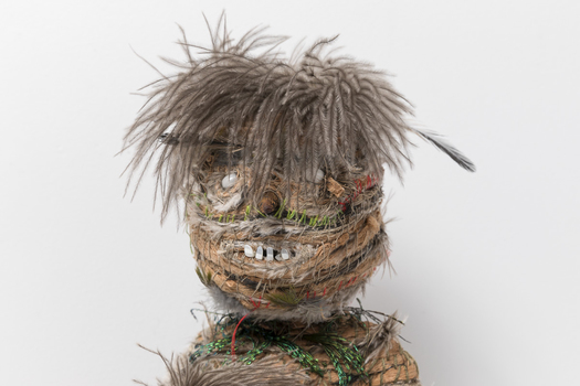 Close up photograph of the head of a woven sculpture. It has rows of feathers and raffia with brown feathers for hair and, seeds for eyes and teeth.
