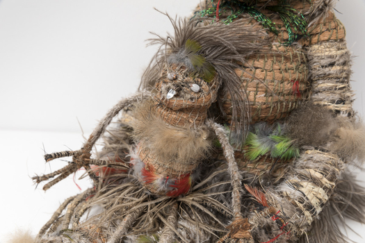 Close up of a small woven figure seated in a larger figure's lap. Made of woven feathers, rafia and seeds for eyes and mouth.