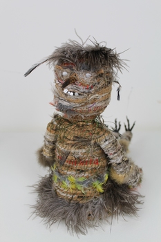 Photograph of the back of the sculpture, showing a figure's back with a face above where the back of a head should be. Figure is rows of woven feathers, rafia, fibres with seeds for teeth and nose.
