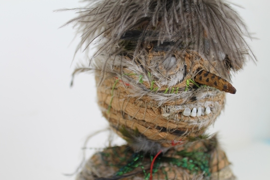 Close up photograph of the head of a woven sculpture. It has rows of feathers and raffia with brown feathers for hair and, seeds for eyes and teeth.