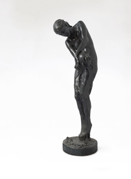 Dark coloured sculpture of a naked woman with a vessel in her hands, from which a liquid pours. The woman's hair is secured in a bun at the back of her head.