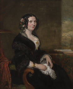 Portrait of a seated woman on wooden chair with green padding and red fabric draped over side, wearing black dress with white lace at bust. She holds white handkerchief in her lap. Her hair is done with ringlets framing her face and flowers adorning side of head. Open book on table behind her