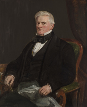 Formal portrait of seated grey haired clean shaven man wearing dark three piece suit. He holds a grey folded object in one hand and glasses in the other. Sits on wooden chair with green padding
