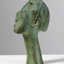 Side angle of a patinated bronze sculpture with a green finish of a whistling man's head. Eyes are slits and nose is elongated.