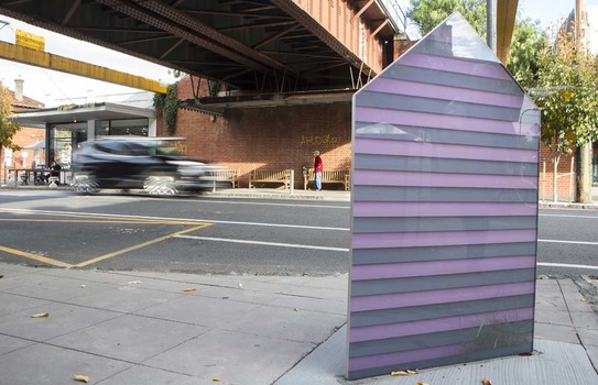 Photograph of an outdoor sculpture in glass in the shape of a bathing box. The bathing box is striped purple and grey. The sculpture is on a footbath below a railway overpass.