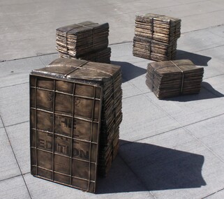 Photograph of an outdoor public art sculpture. The work is comprised of four sculptural stacks of newspapers, cast in bronze, which sit on a concrete footpath.