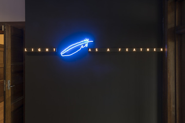 Photograph of a dark wall with a blue and white light artwork installed on it that reads Light as a feather and has a light in the shape of a feather in the centre.