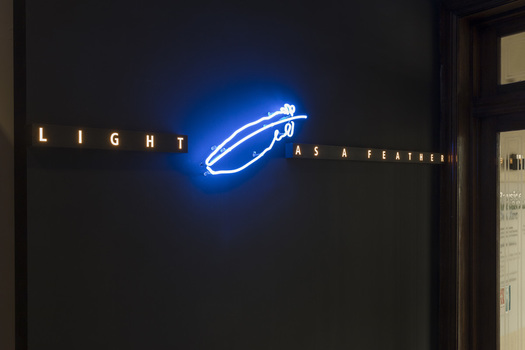 Angled view of a dark wall with a blue and white light artwork installed on it that reads Light as a feather and has a light in the shape of a feather in the centre.