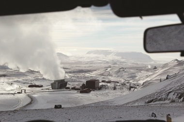 Icelandic snow covered landscape viewed through the front window of a car. Power plant buildings at centre with large plume of smoke or steam emitting towards left from one of the buildings. Mountains partially obscured by cloud in background.