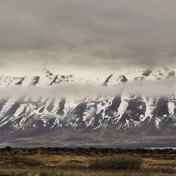 Colour photograph of an Icelandic landscape partially with grey cloudy sky. Snow covered mountains partially obscured by cloud in centre. Brown grasses and low vegetation in foreground.