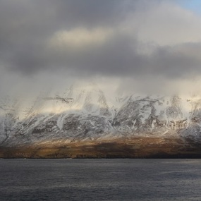 Colour photograph of an Icelandic landscape partially with grey cloudy sky. Snow covered mountains partially obscured by cloud in centre. Brown grasses and low vegetation in foreground.