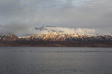 Colour photograph of an Icelandic landscape of snow covered mountains partially obscured by cloud in centre and with sun shining on the upper parts of the mountain. Body of water in foreground. Grey cloudy sky with glimpse of blue at the top.