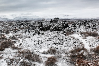 Colour photograph of an Icelandic landscape partially covered in snow. Nude bushes and trees in foreground, partially obscuring a snow covered path or road. Rocky outcrop or hill with dark stones in centre. Mountains in background partially obscured by cloud. Grey cloudy sky.