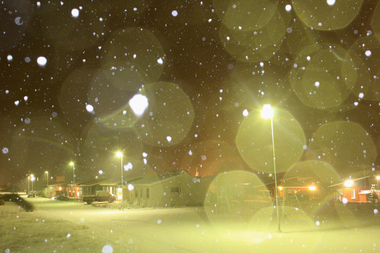 Colour photograph of celandic landscape of snow covered street, houses, street lamps and cars at night. Street lamps bathe snow and houses in a yellow light. Sky is dark with snow flakes falling. Snow and light create semi circular patterns across entire surface of photograph.