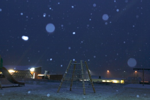 Colour photograph of Icelandic landscape of snow covered playground at night. Buildings with lights on the outside or shining through windows in centre. Sky and snow appears blue. Snowflakes are falling and some appear as circular shapes across the photograph.