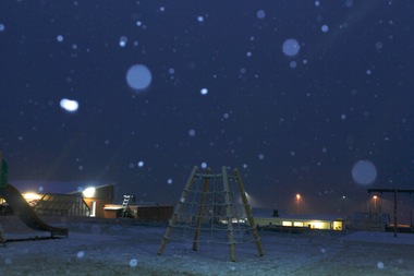 Colour photograph of Icelandic landscape of snow covered playground at night. Buildings with lights on the outside or shining through windows in centre. Sky and snow appears blue. Snowflakes are falling and some appear as circular shapes across the photograph.