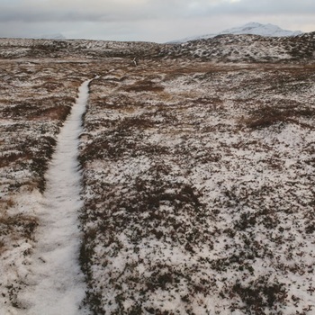 Colour photograph of  Icelandic snow covered landscape with brown low vegetation. A white path runs from lower left corner along right side and up towards centre. Hills and mountains in background. Grey cloudy sky.