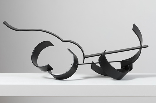 Photograph of a black, welded steel abstract sculpture sitting on a white plinth. Geometric shapes are adjoined to form cluster in roughly horizontal orientation.