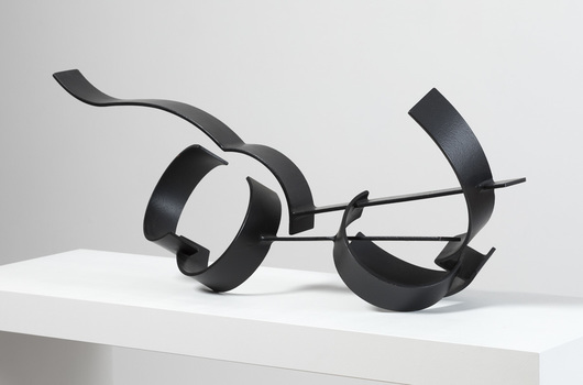 Photograph of a black, welded steel abstract sculpture sitting on a white plinth. Geometric shapes are adjoined to form cluster in roughly horizontal orientation.