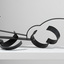 Photograph of a black, welded steel abstract sculpture sitting on a white plinth. Geometric shapes are adjoined to form cluster in roughly horizontal orientation.