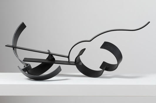 Photograph of a black, welded steel abstract sculpture sitting on a white plinth. Geometric shapes are adjoined to form cluster in roughly horizontal orientation.