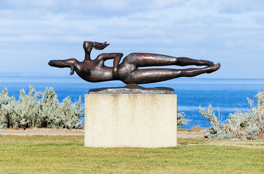 Cast bronze sculpture of a female figure, reclining on its right side, with elbows at shoulder height and her legs kicking, as if to indicate that she is swimming. Her hair appears to float behind her. The figure is attached to a low bronze base at the hip. The sculpture sits on a rectangular cement plinth 