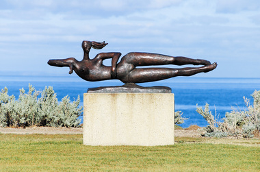 Cast bronze sculpture of a female figure, reclining on its right side, with elbows at shoulder height and her legs kicking, as if to indicate that she is swimming. Her hair appears to float behind her. The figure is attached to a low bronze base at the hip. The sculpture sits on a rectangular cement plinth 