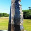 Photograph of an outdoor marble sculpture, abstract shape in marble, ship rudder with sand castle shape at top. Green vegetation is visible behind the sculpture.
