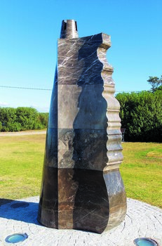 Photograph of an outdoor marble sculpture, abstract shape in marble, ship rudder with sand castle shape at top. Green vegetation is visible behind the sculpture.