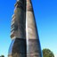 Photograph of an outdoor marble sculpture taken from a low vantage point. The sculpture is an abstract shape in marble, ship rudder with sand castle shape at top. Green vegetation is visible behind the sculpture.