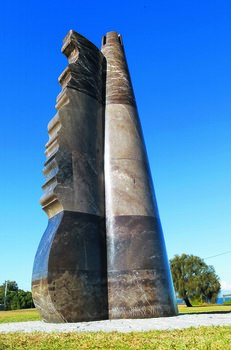 Photograph of an outdoor marble sculpture taken from a low vantage point. The sculpture is an abstract shape in marble, ship rudder with sand castle shape at top. Green vegetation is visible behind the sculpture.